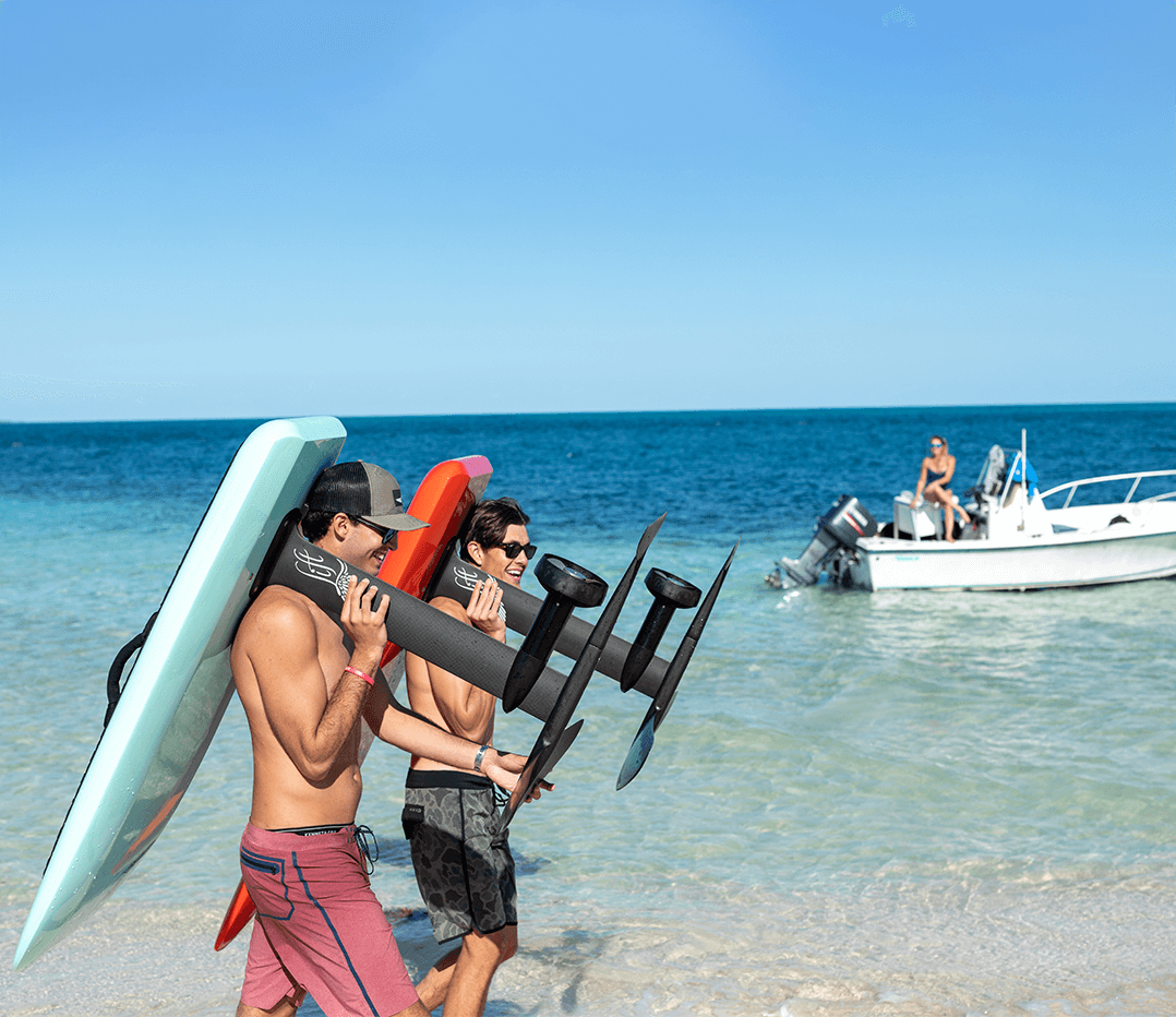 Two men carrying surfboards to the water on a sunny beach day, with a boat in the background.